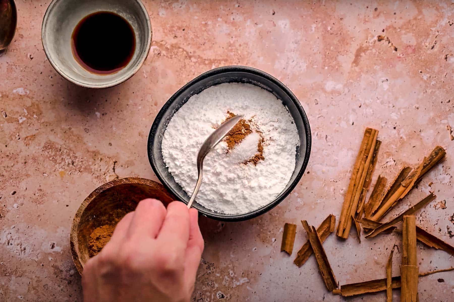 A hand uses a spoon to mix cinnamon into a bowl of powder, surrounded by sticks of cinnamon and a bowl of cinnamon glaze on a textured surface.