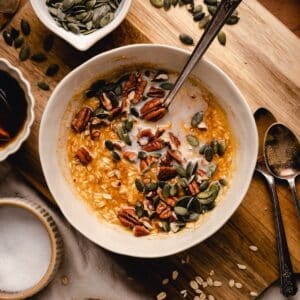 A bowl of pumpkin oatmeal topped with pecans and pumpkin seeds rests on a wooden surface, surrounded by spoons and a small dish of seeds.