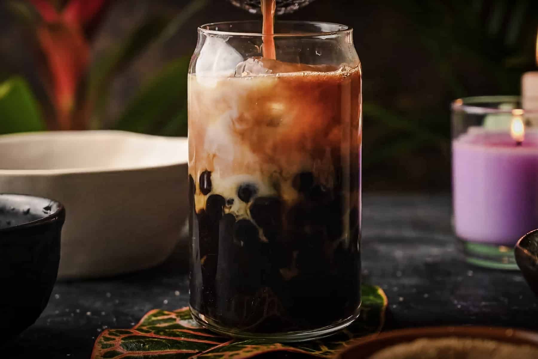 A glass of iced bubble tea with milk cascading in, set among a flickering candle and assorted bowls on a dark surface.
