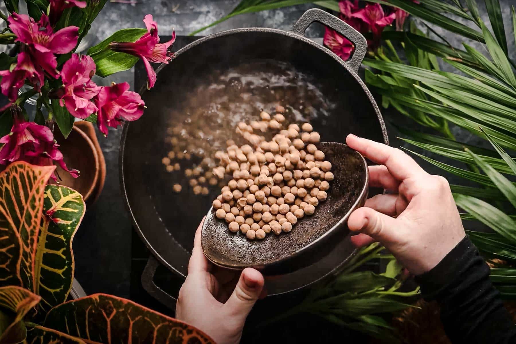 A person pouring tapioca pearls into a large pot, surrounded by pink flowers and lush green foliage.