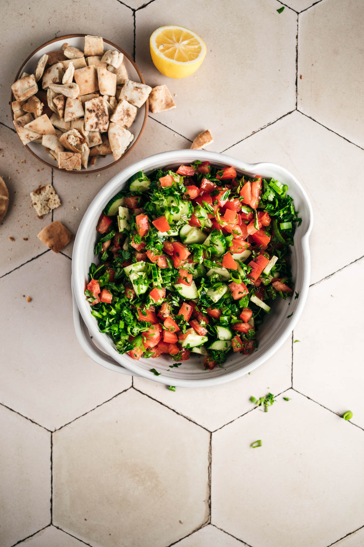 A bowl of chopped vegetable salad with tomatoes, cucumbers, and greens on a tiled surface. A plate of pita bread and a half lemon are placed beside the bowl.
