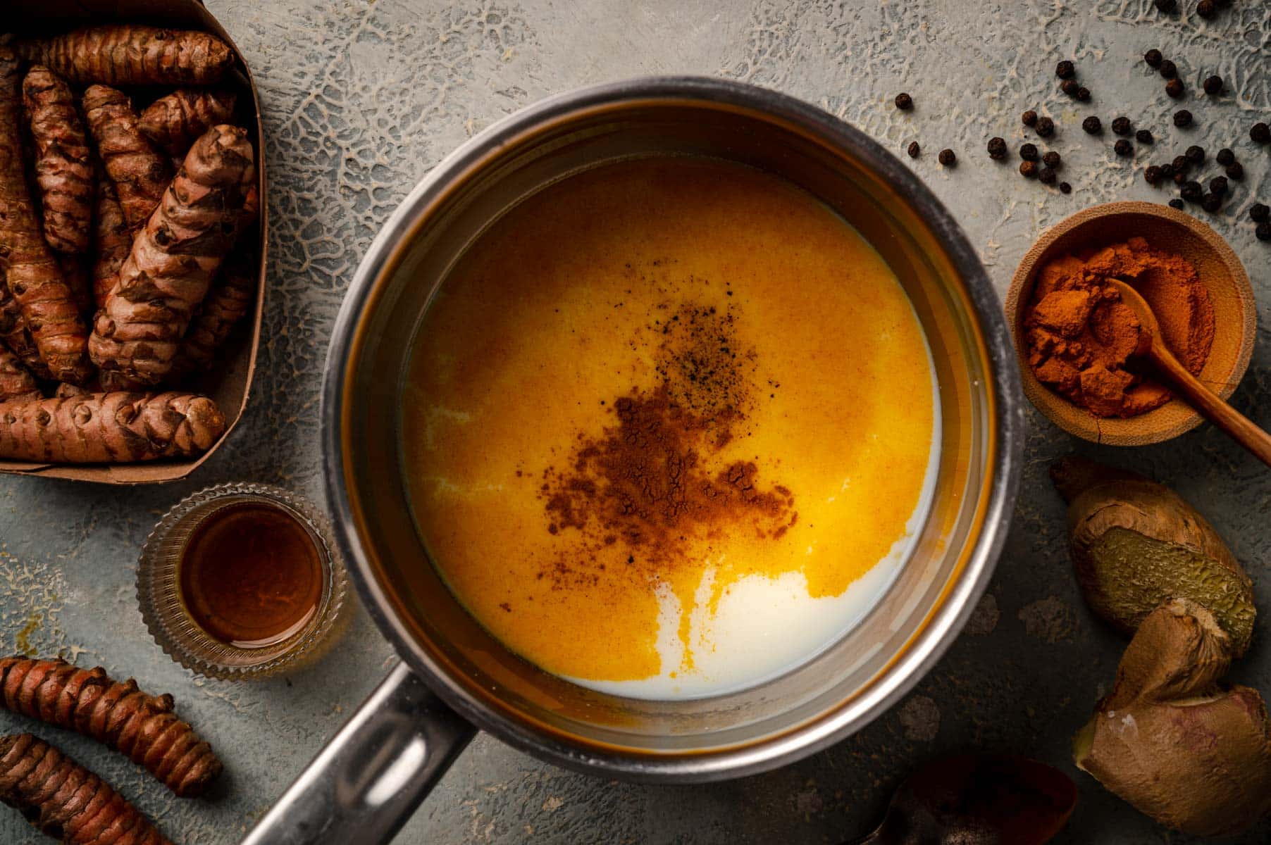 A pot containing a mixture of almond milk and turmeric is surrounded by fresh turmeric roots, ground turmeric in a bowl, black peppercorns, and a small glass of honey on a textured surface.