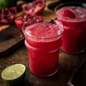 Two glasses of pomegranate limeade with ice sit on a wooden table.