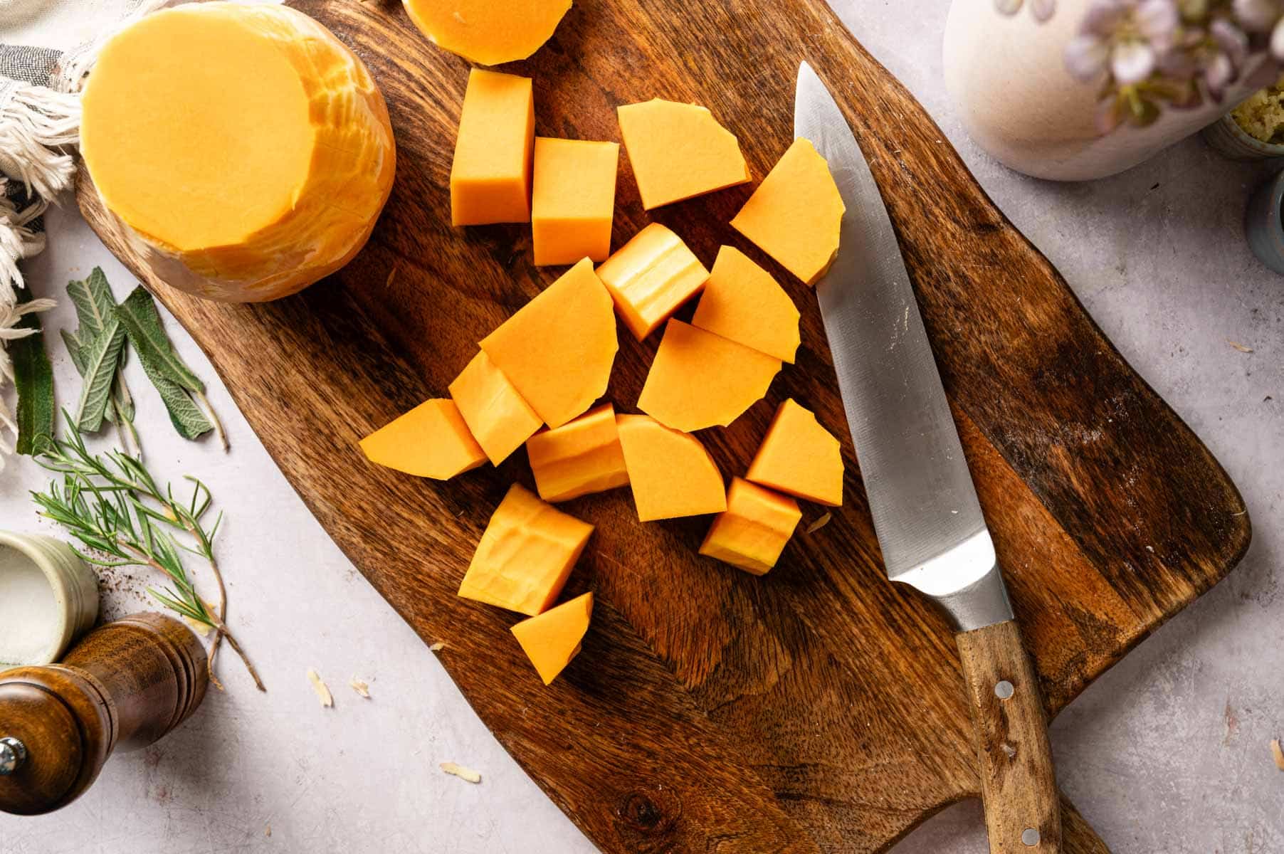Butternut squash on a wooden cutting board being peeled and then chopped into cubes.